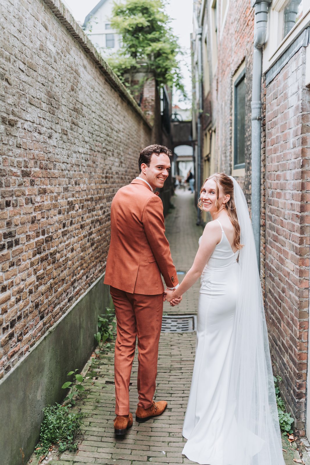 Bruidspaar hand in hand in een steegje, de bruid kijkt lachend om
