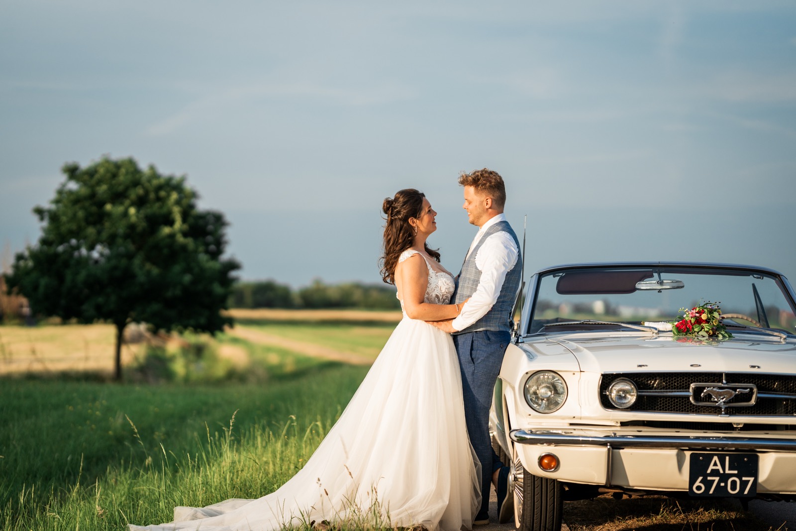 Bruidspaar bij een witte Mustang in het weiland bij zonsondergang