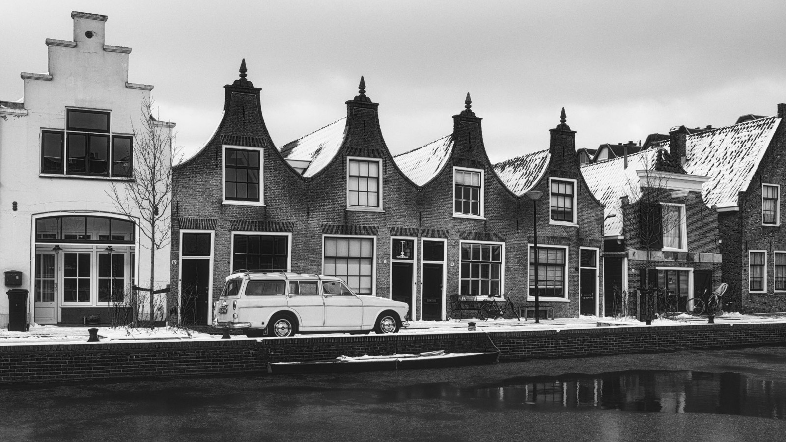 Zwart-witfoto van oude grachtenpanden met klokgevels en een klassieke auto in de sneeuw